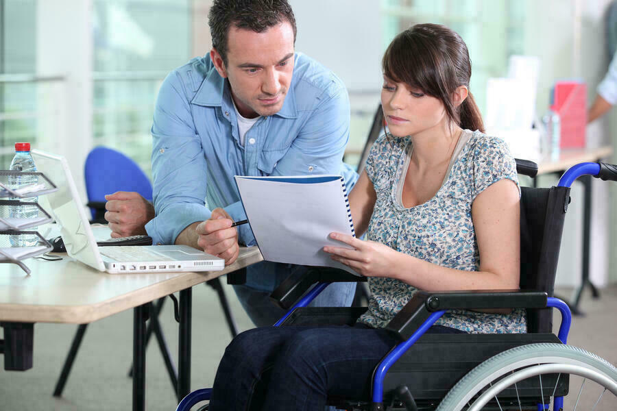 Eine Frau im Rollstuhl und ein Kollege besprechen Notizen an einem Schreibtisch.