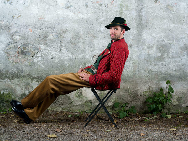 Mann in roter Trachtenjacke mit Hut sitzt auf einem Hocker und hat die Hände auf den Schoß gelegt. Er hat die Beine übereinander gekreuzt und schaut in die Kamera.