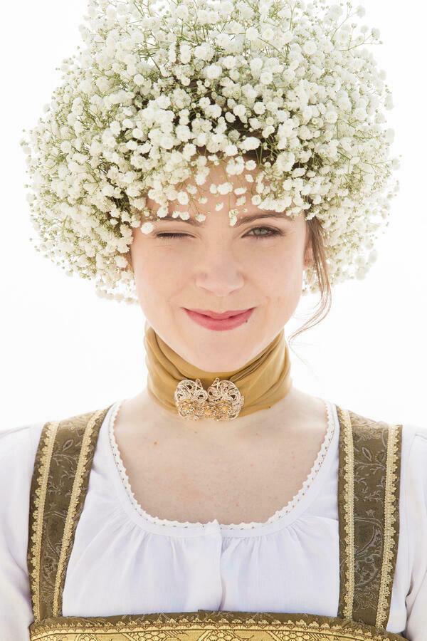 Eine Frau mit Dirndl und Halstuch mit Brosche schaut in die Kamera. Sie hat weiße Blumen in der Frisur und zwinkert mit dem rechten Auge.