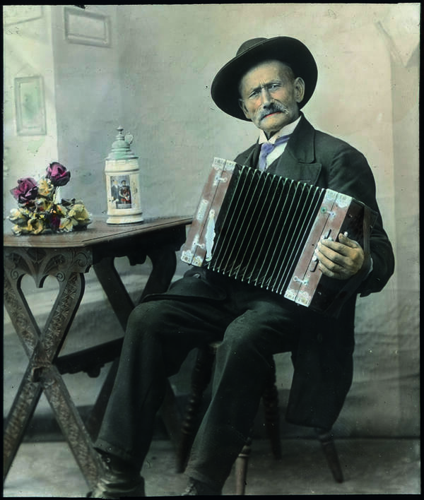 Auf dem handkolorierten Foto sieht man einem Mann mit Hut und Akkoerdeon, der auf einem Wirtshausstuhl sitzt und spielt. Es handelt sich um Max Wörl auf einer Fotografie von Georg Pettenkofer. 