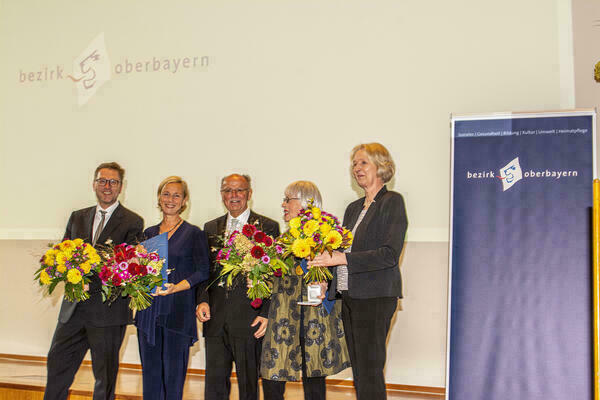 Gruppenfoto mit Blumen vor einer Projektionswand mit dem Logo des Bezirks Oberbayern.