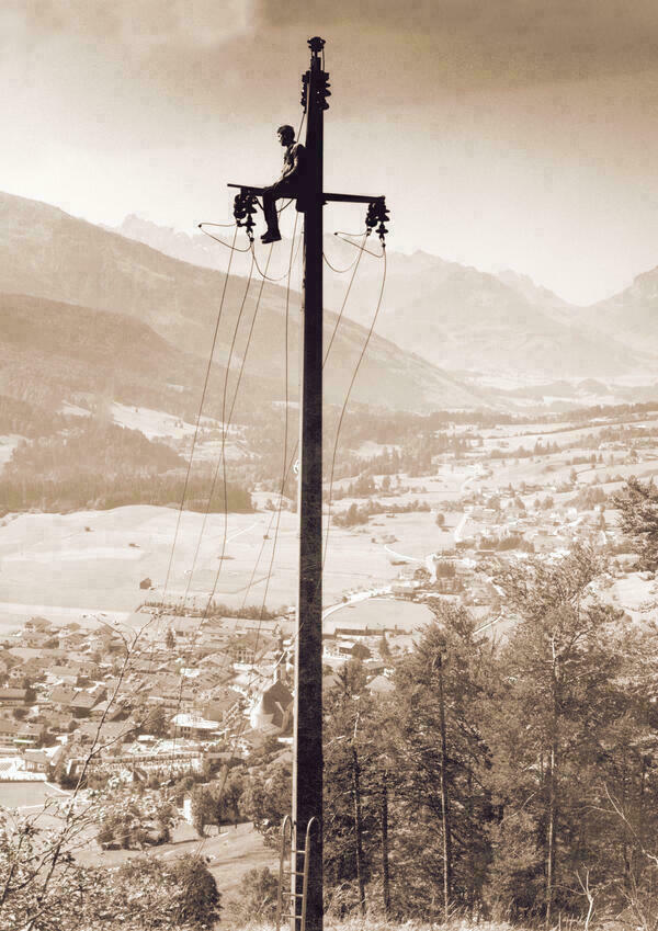 Arbeiter auf einem Hochspannungsmasten hoch über einem Dorf vor der Kulisse der Alpen.