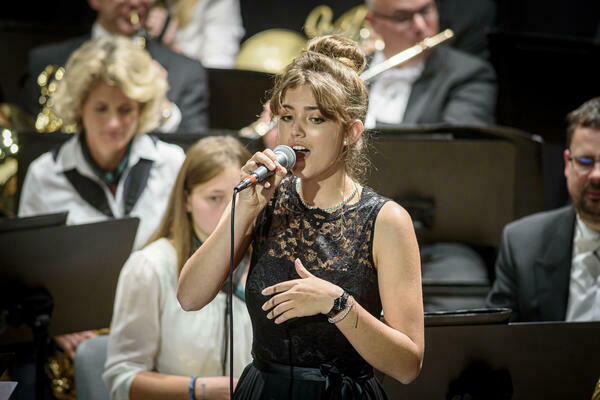 Eine junge Frau ist in der BIldmitte zu sehen; sie singt in ein Mikrofon. Im Hintergrund erahnt man ein Orchester.