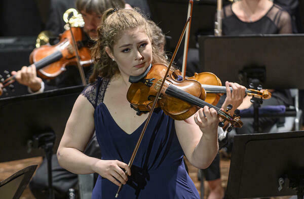 Eine junge Frau mit einem Streichinstrument ist in der Bidmitte zu sehen, im Hintergrund erahnt man ein Orchester.