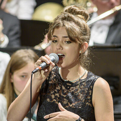 Eine junge Frau ist in der BIldmitte zu sehen; sie singt in ein Mikrofon. Im Hintergrund erahnt man ein Orchester.