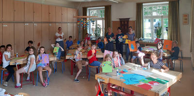 Kinder und Lehrerinnen im Werkraum der Schule. Auf den Tischen siehrt man bunte Papierbögen