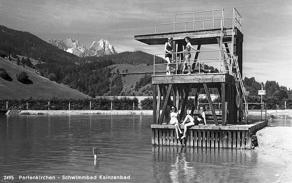 Schwarz-Weiss-Aufnahme eines Sprungturms in einem Freibad. Am Turm stehen und sitzen mehrere Menschen. Im HIntergrund ist Alpenpanaorama zu sehen.