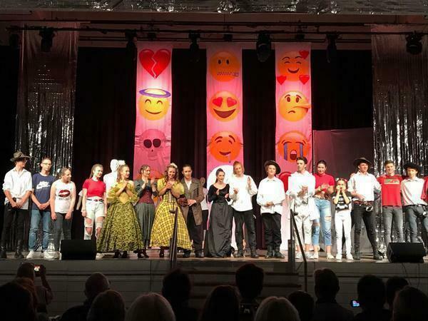 Kostümierte Menschen stehen nebeneinander in einer Reihe auf einer Bühne, im Hintergrund hängen Banner mit verschiedenen Emojis