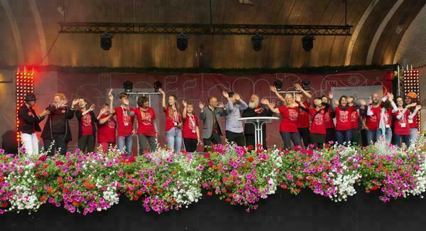 Viele Menschen stehen ein einer Reihe auf einer Bühne; die meisten haben die Hände winkend in der Luft. Viele tragen ein rotes T-Shirt mit dem Aufdruck 