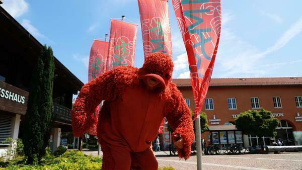 Ein kostümierter Schauspieler posiert vor dem Kongresshaus Garmisch-Partenkirchen. Hinter ihm eine Reihe hoher Fahnen mit dem Aufdruck 