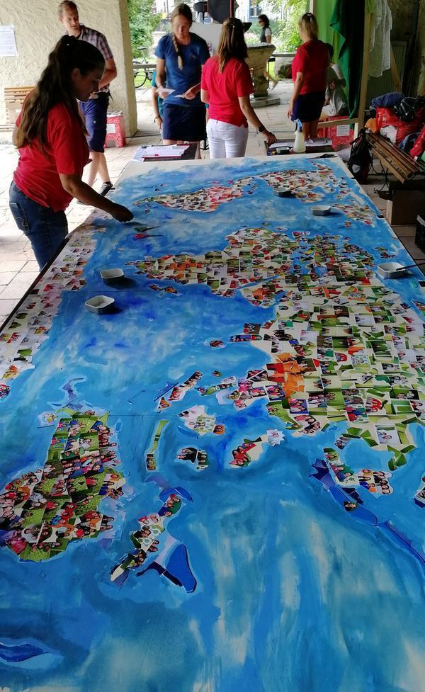 Auf einem großen Tisch liegt eine gemalte Weltkarte. Auf diese wurden viele Fotos von Menschen geklebt. Um den Tisch herum stehen mehrere Jugendliche. 
