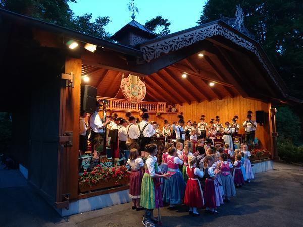 Auf einer Bühne ist eine in bayerischer Tracht gekleidete Kapelle zu sehen, vor der Bühne sind Kinder, ebenfalls in bayerischer Tracht gekleidet. 