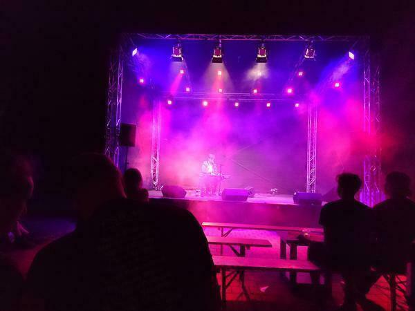 Auf einer violett ausgeleuchteten Bühne ist ein Musiker an einem Keyboard zu erkennen. Im Vordergrund sind die die dunklen Silhouetten von Zuhörern zu sehen. 
