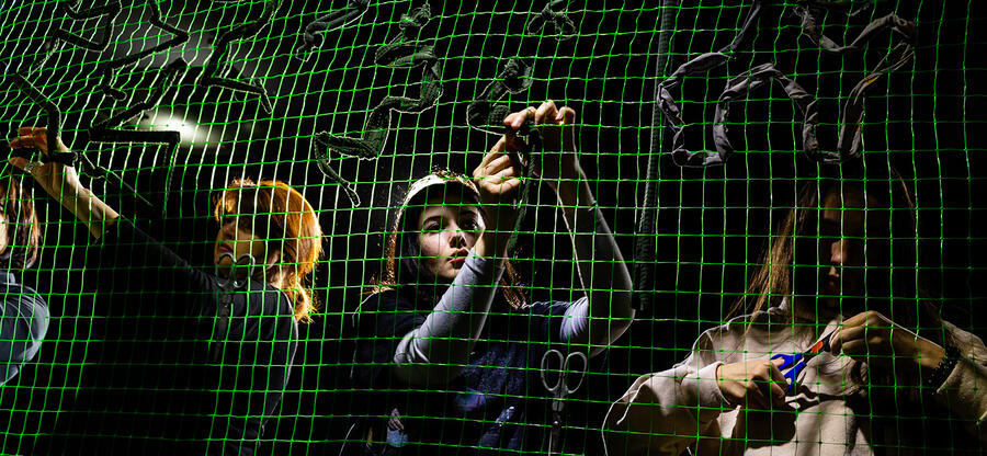The Thin Line / Der schmale Grat - Ausstellung mit ukrainischen Fotografien; Bild: Freiwillige beim Knüpfen von Tarnnetzen, junge Frauen im Scheinwerferlicht hinter einem grünen Netz; Fotografin: Kateryna Moskalyuk