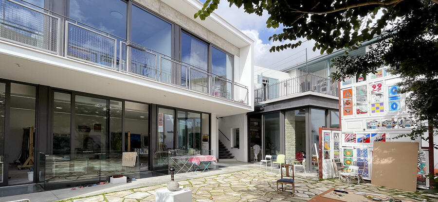 Innenhof der MeMeraki Artist Residency in Limassol; Bild: modernes Ateliergebäude mit Glasfront links, Natursteinpflaster in der Mitte vorne, Bilderwand rechts; sonnige helle Anmutung