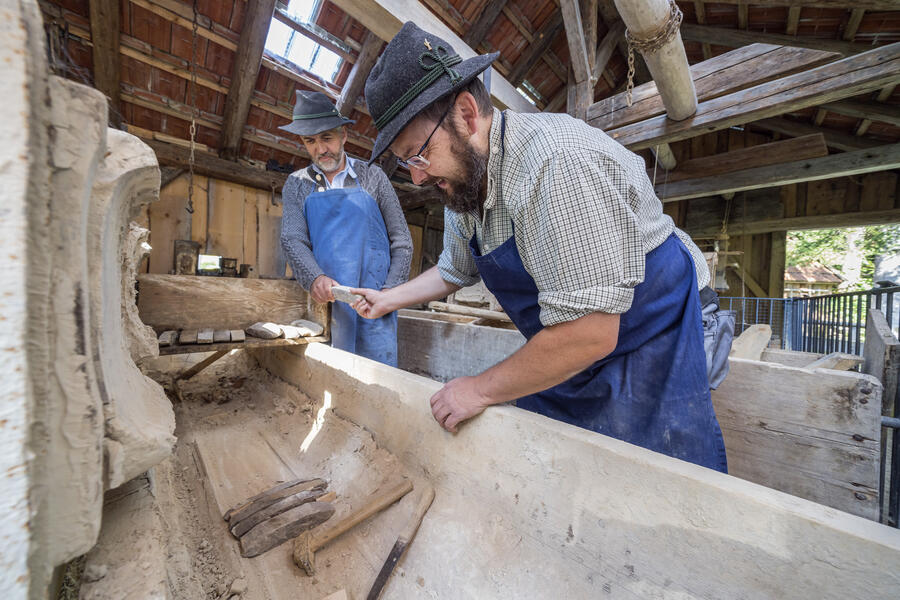 Zwei Männer in Arbeitskleidung richten gemeinsam kleine, geformte Steine in einen Trog aus Holz ein.