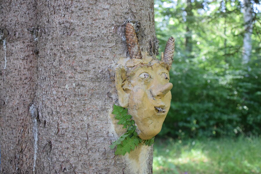 An einem Baumstamm hängt eine Maske aus Ton, die mit Naturmaterialien verziert ist.
