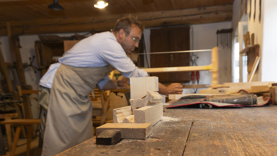 Auf einer Werkbank ist ein Stück helles Holz eingespannt. Im Hintergrund arbeitet ein Mann mit einer Säge.