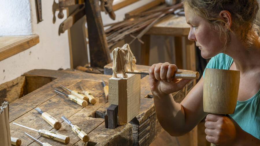 Eine junge Frau bearbeitet mit Werkzeugen ein Stück Holz. Sie schnitzt daraus eine Kuh.