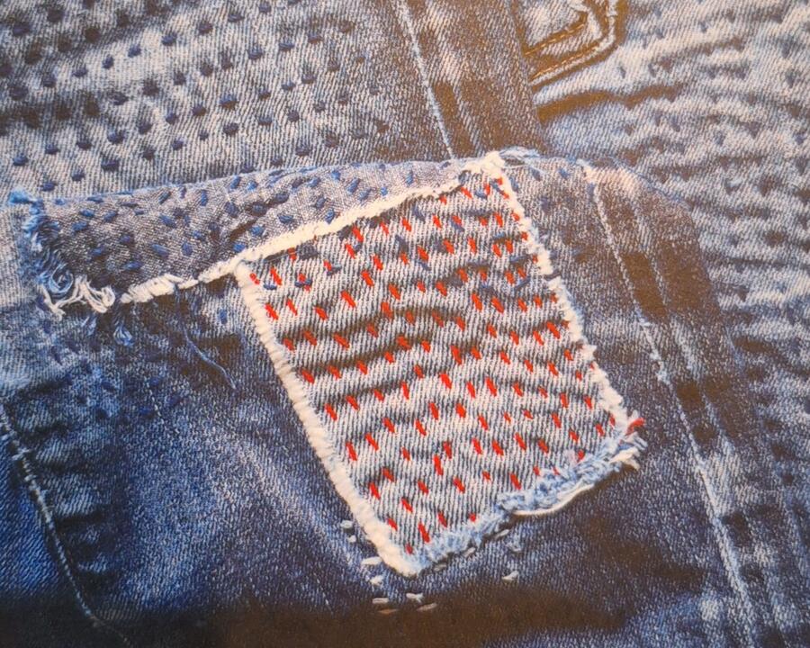 Auf einem Jeansstoff ist mit rotem Faden ein Flicken aufgenäht und gestickt.