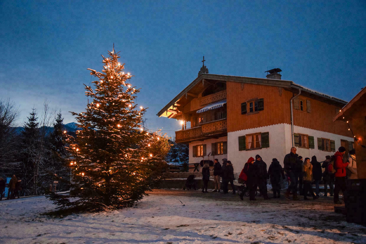 Ein leicht schneebedeckter Weihnachtsbaum mit Lichtern neben einem Haus, davor eine kleine Menschenmenge.