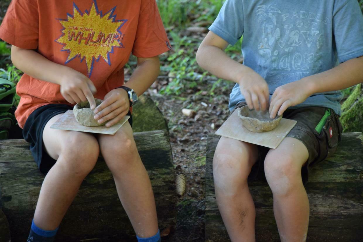 Zwei Kinder sitzend, auf ihrem Schoß jeweils eine Schüssel aus Ton, die sie mit ihren Händen bearbeiten