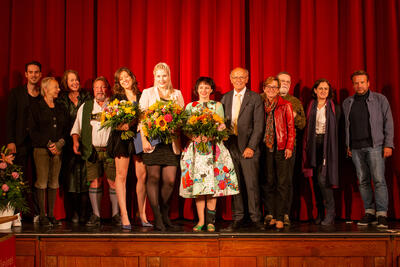 Eine Gruppe von sieben Frauen und fünf Männern steht auf einer Bühne vor rotem Vorhang. Drei Frauen halten Blumensträuße.