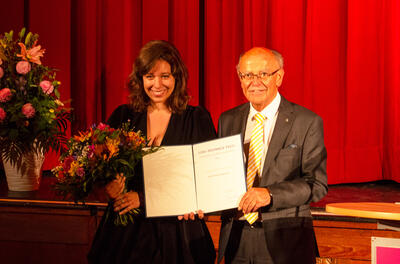 Eine junge Frau mit Blumenstrauß steht neben einem älteren Mann im Anzug, der eien Urkunde hält. Im Hintergrund ein roter Bühnenvorhang.