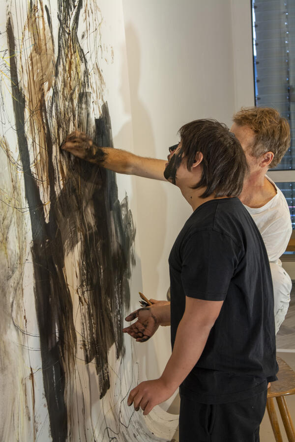Zwei Männer malen auf einer großen Leinwand mit ihren Händen ein abstraktes Kunstwerk aus grauen, braunen und schwarzen Strichen.