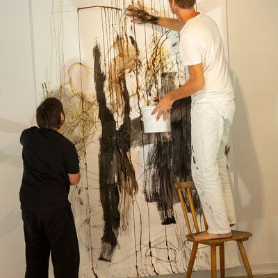 Ein Mann in schwarzer Kleidung steht neben einem Mann in Weiß.  Der steht euf einer Leiter und bearbeitert eine große Zeichnung an der Wand mit seinen Händen.