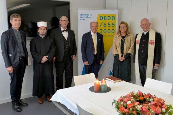 Ein Foto von sechs Personen, die sich in einem Raum des Krisendienstes Psychiatrie Oberbayern befinden. Vor Ihnen ist ein Tisch mit einer weißen Tischdecke, vier Kerzen und Blumen zusehen.