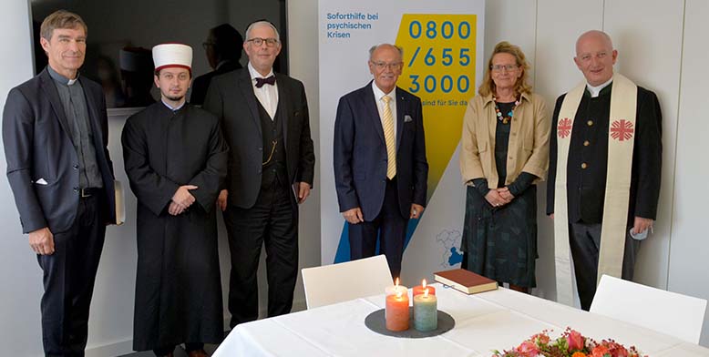 Ein Foto von sechs Personen, die sich in einem Raum des Krisendienstes Psychiatrie Oberbayern befinden. Vor Ihnen ist ein Tisch mit einer weien Tischdecke, vier Kerzen und Blumen zusehen.