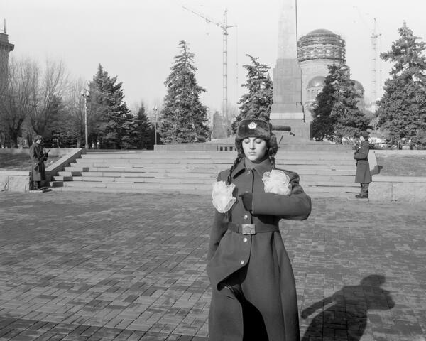 Kunstwerk Fotografie: Eine junge Frau in osteuropäischem Militätmantel und Fellmütze patroulliert vor einem Denkmal. Ihre Haare sind zu Zöpfen geflochten, an denen große weiße Puscheln hängen.