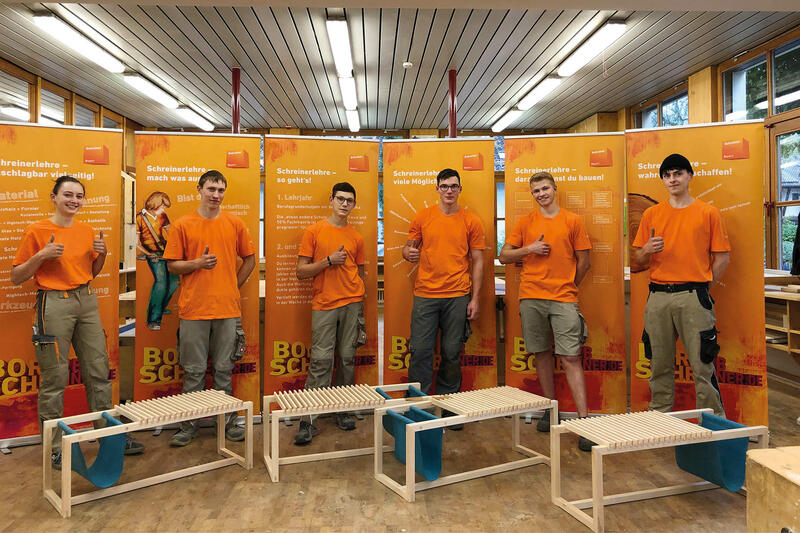 Sechs junge Männer mit orangenem T-Shirt stehen vor sechs orangenen Rollups. Vor jedem Mann steht ein selbsgeschreinertes Möbelstück.