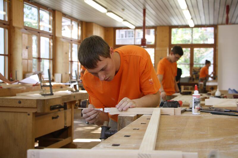 Ein junger Mann in orangenem T-Shirt arbeitet in einer Schreinerei an einem Mbelstck.
