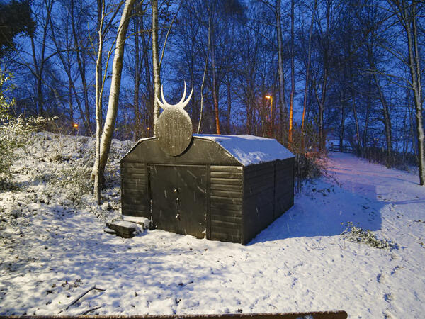 In einer verschneiten Landschaft mit lichtem Baumbestand ist als Kunstobjekt ein schwarzes Gartenhaus mit stilisiertem Ochsenkopf zu sehen.
