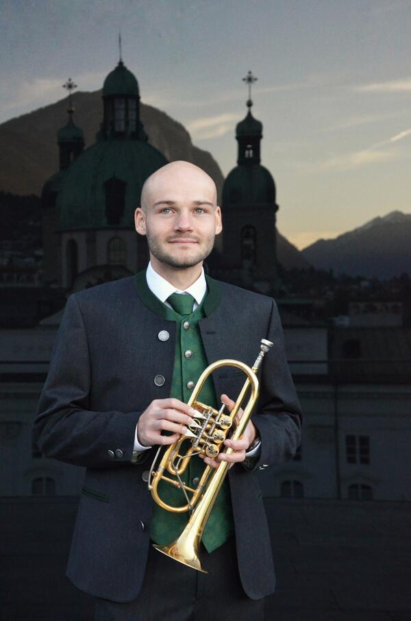 Bernhard Achhorner mit Trompete in traditioneller Kleidung. Im Hintergrund abendliches Alpenpanorama und eine Kirche.