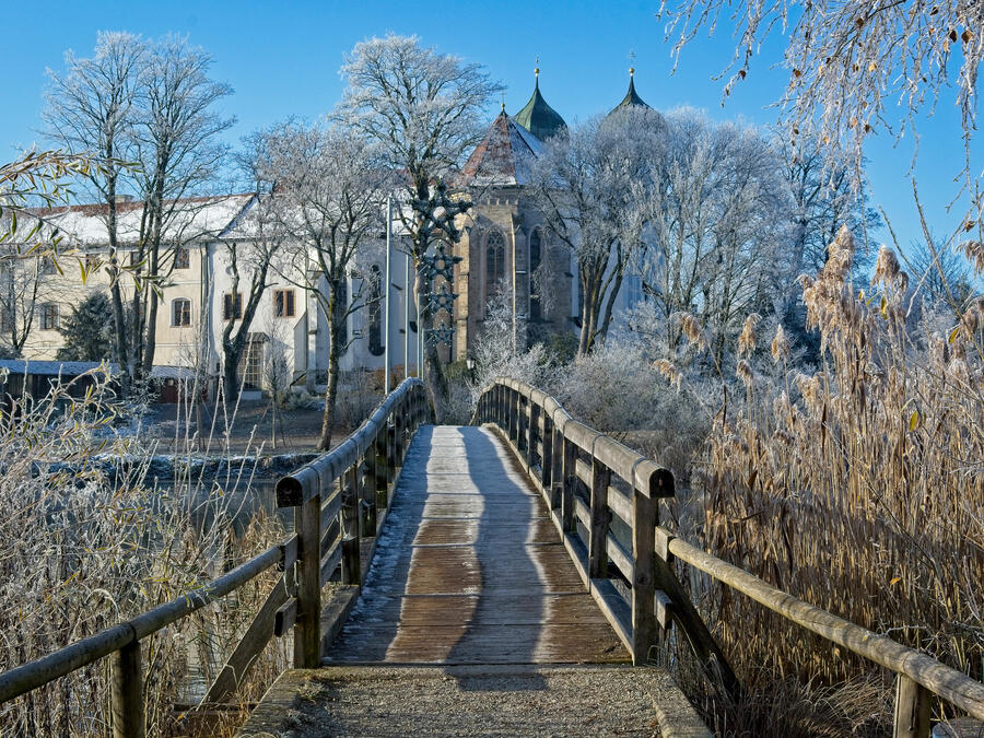 Eine leicht schneebedeckte Holzbrücke führt über einen See, hin zum von kahlen Bäumen verdeckten Kloster Seeon.