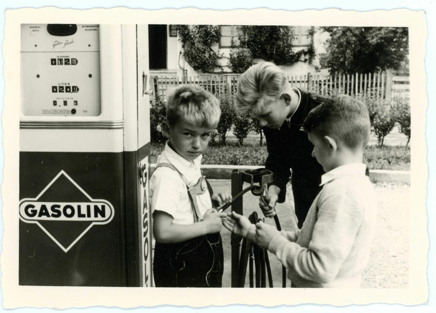 Ein altes schwarz-weiß Bild mit drei Jungs an einer Tankstelle. Einer von ihnen, gekleidet in bayrischer Tracht, lehnt an der Zapfsäule. Auf ihr die Aufschrift "Gasolin".