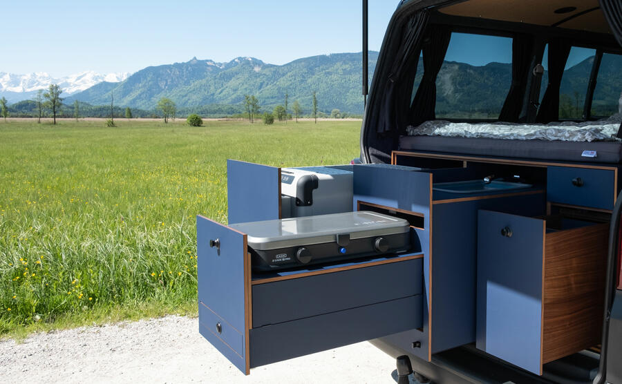 Aus dem Kofferraum eines VW-Bus ragt ein Schubladensystem aus blau lackiertem Holz. Im Hintergrund eine saftig grüne Wiese mit Bergen.