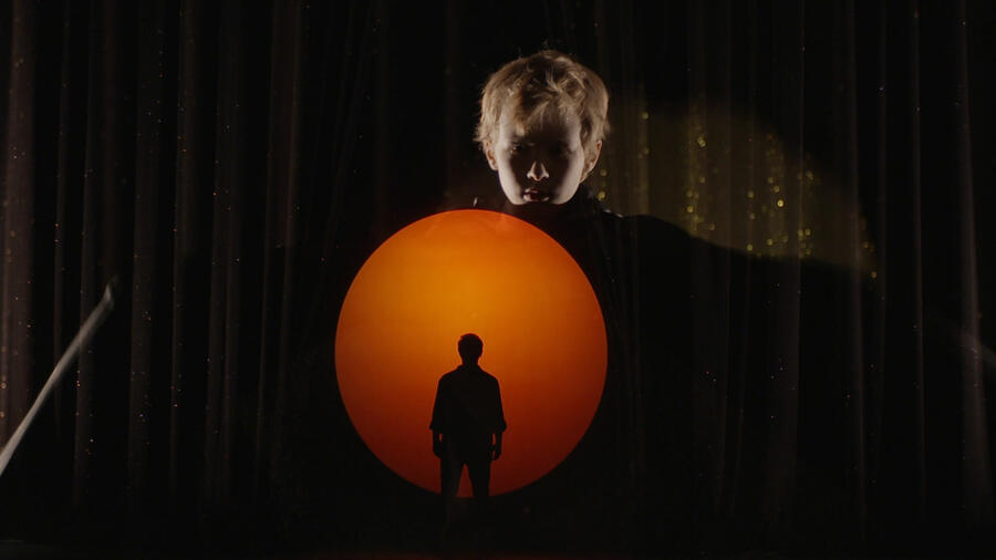 Bühnenbild einer Aufführung: Die Silhouette einer Person vor einer orangenen Abendsonne. Im Hintergrund wird ein blonder Mann an den Vorhang projiziert.
