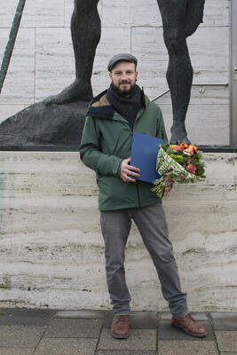 Mann mit Jacke und Schiebermütze steht mit einem Blumenstrauß vor einer Bronzefigur. Von der Statue erkennt man nur die Füße.