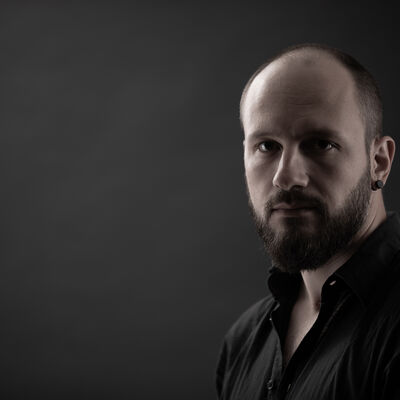 Porträtfoto: Mann mit Bart und kurzen Haaren vor dunklem Hintergrund