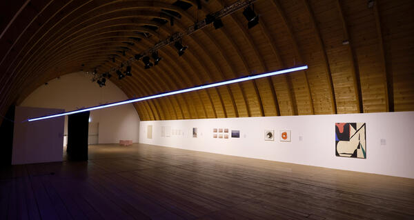 Blick in einen Ausstellungsraum: An den Wänden hängen Gemälde, in der Raummittel hängt ein Leuchtband als Teil einer Kunstinstallation.