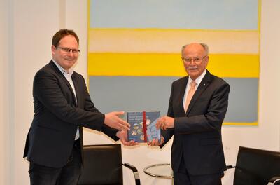 Bezirkstagspräsident Josef Mederer überreicht einem Herrn mit Brille ein Buch.