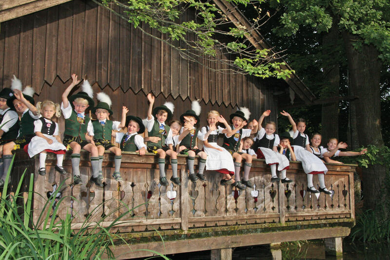 Ausschnitt eines Holzstadels mit verziertem Balkon, auf dem Kinder in Tracht sitzen und winken