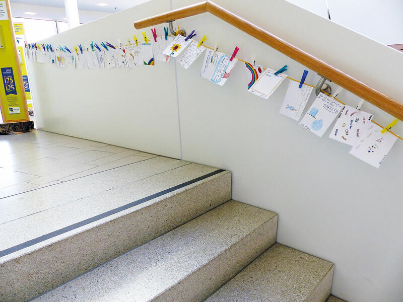 Treppengeländer mit einer parallel laufenden Schnur, an der Kinderzeichnungen befestigt sind.