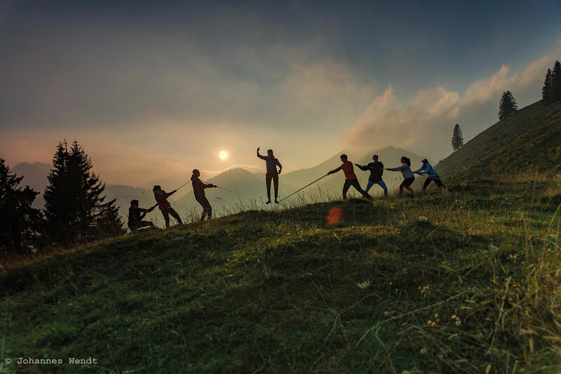 Bergwiese bei Sonnenuntergang mit der Silhouette von acht Personen; eine Person steht auf einer Slackline, die anderen halten das Seil.