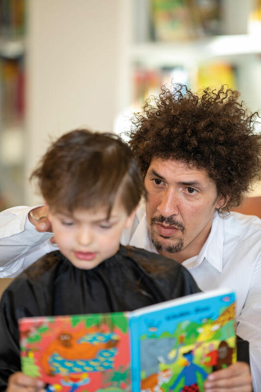 Ein Mann mit dunklen Afrohaaren und weißem Hemd steht hinter einem Jungen mit schwarzem Friseurumhang und schneidet ihm die Haare. Der Junge hält ein Kinderbuch in der Hand.