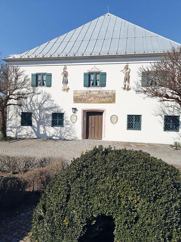 Vorderansicht eines zweistöckigen Gebäudes mit weißer Fassade mit Lüftlmalerei und hellgrünem spitzen Dach 